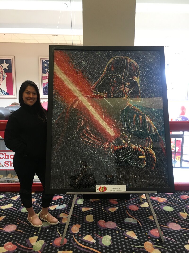 Kathleen stands next to a large framed portrait of Darth Vader made of Jelly Beans. She is wearing all black and white shoes. She has long dark hair and is smiling.