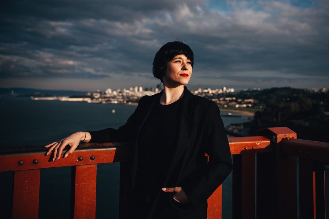 Natalie stands on the Golden Gate Bridge, looking off camera. She has light skin, is wearing all black and a hat, and is wearing red lipstick.