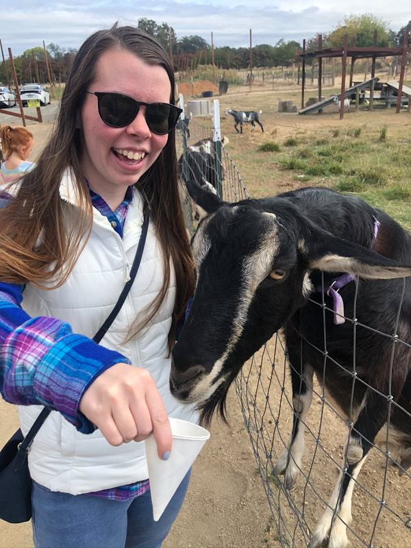 Andrea Northrup smiles as a goat eats out of her hand. She is a white woman with long brown hair, and is wearing sunglasses and a vest under a flannel.
