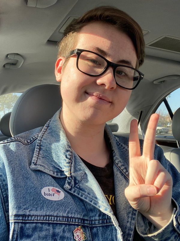Nathan Burns smiles and makes a peace sign at the camera. They are a white person with short blonde hair and are wearing glasses. They are wearing a jean jacket with an 