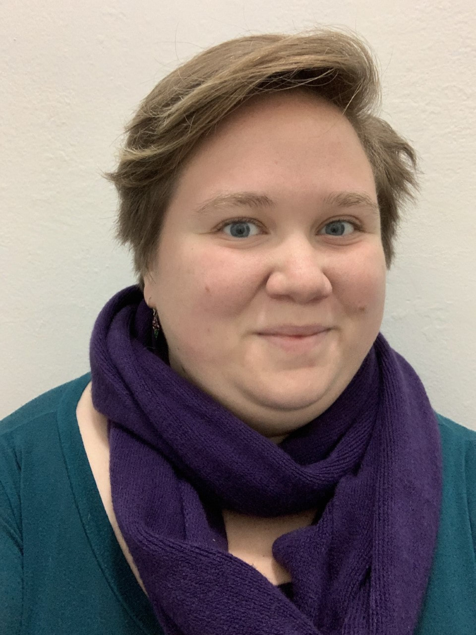 Azaleah, a white cisgender woman with short brown hair, wearing a purple scarf and a teal shirt, smiles confidently at the camera.